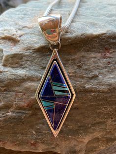 This beautifully well crafted Diamond shape Pendant has been handmade by a Navajo Artisan. The Colors of the Pendant come from Lapis Lazuli, Turquoise and Blue Opal. The Pendant measures 2.25” long and 3/4” wide. There are two pendants available and the design will vary. “Winds of Wisdom” (Lapis Lazuli, Turquoise and Blue Opal. The Navajo (Dine’) believe that the wind is a powerful source. The native tradition believes that at any moment, the blowing wind can inspire one with hope, resolve, and Blue Sterling Silver Necklace With Inlay, Unique Blue Teardrop Turquoise Necklace, Blue Southwestern Necklace With Large Pendant, Southwestern Style Blue Necklace With Large Pendant, Traditional Turquoise Inlay Necklace As Gift, Traditional Turquoise Necklace With Inlay For Gift, Artisan Blue Inlay Necklaces, Southwestern Blue Teardrop Jewelry, Unique Blue Necklace With Inlay