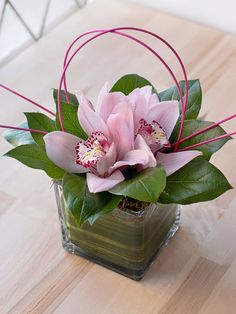 a pink flower arrangement in a square glass vase on a wooden table with greenery