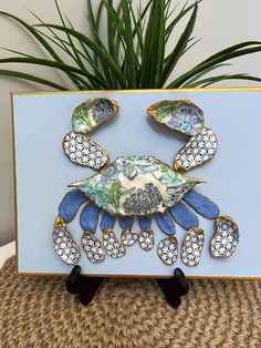 a blue and white crab with gold accents on it's back, sitting next to a potted plant