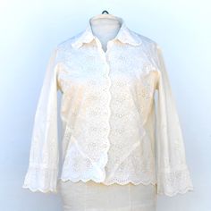 This beautiful Saks Fifth Avenue cotton blouse has an elaborate  eyelet and embroidered design. It is is very good condition and show little sign of wear, but the yellow embroidered tag is a fraying at the ends and the blouse may have a general age-related discolouration that makes it more of an ivory colour and not a stark white. The front has plastic shank style buttons up the front and there are bust darts. Around the bottom of the front are 2 diagonal darts, as well.  The design of the embro Elegant Cotton Eyelet Tops, Cotton Eyelet Long Sleeve Tops, Elegant Long Sleeve Cutwork Top, Spring Cotton Blouse With Lace Work, Spring Cotton Lace Work Blouse, White Cotton Shirt With Chikankari Embroidery, White Cotton Chikankari Embroidered Shirt, Fitted Cotton Blouse With Broderie Anglaise, Traditional Cream Cotton Top