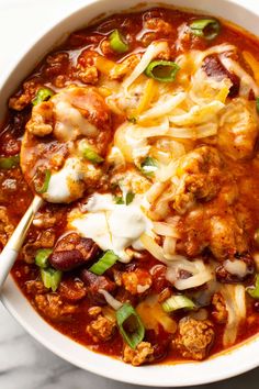 a white bowl filled with chili and meat covered in cheese, sour cream and green onions