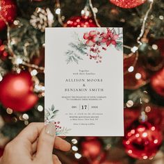 a person holding up a wedding card in front of a christmas tree with red and green ornaments