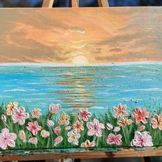 an easel with flowers painted on it in front of the ocean and sun setting