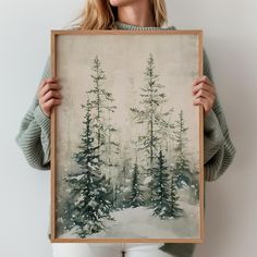 a woman holding up a painting with pine trees in the background and snow on the ground