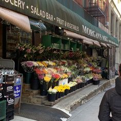 a woman walking past a flower shop with lots of flowers on the side of the street