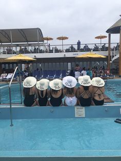 four women wearing hats sitting on the edge of a swimming pool