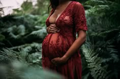 a pregnant woman in a red dress is posing for the camera while surrounded by ferns