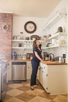 a woman standing in a kitchen reaching for something on the shelf above her head with one hand