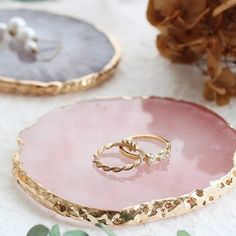 two gold wedding rings sitting on top of a pink agate stone tray next to pine cones