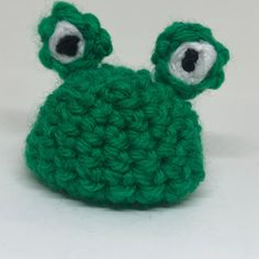 a green crocheted frog with big eyes on it's head, sitting in front of a white background