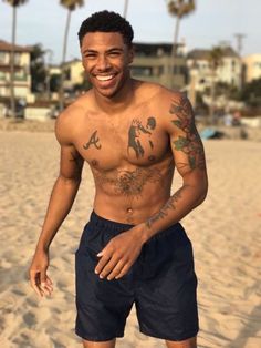 a shirtless man standing on the beach with his hands in his pockets and smiling