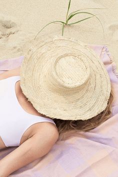 Woven hat on top of model's head that's laying down with a pink and purple fabric on the beach. Summer Fedora Panama Hat Made Of Palm Leaf, Beachy Straw Hat For Travel, Spring Beachwear Sun Hat In Natural Color, Natural Color Beachwear Sun Hat For Beach Season, Summer Beachwear Straw Hat For Travel, Natural Color Beachwear Sun Hat For Spring, Summer Beachwear Sun Hat In Toquilla Straw, Beachwear Straw Hat For Vacation Travel, Beachwear Straw Hat For Travel And Vacation