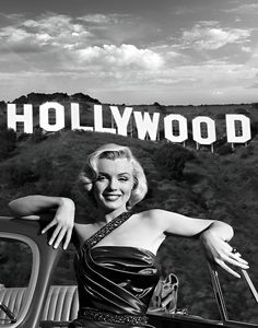 a black and white photo of a woman leaning on the back of a car with hollywood sign in the background