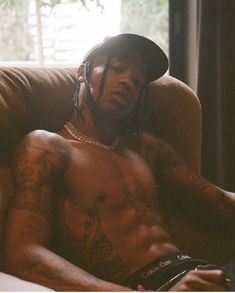 a shirtless man sitting in a chair with tattoos on his chest and arms, wearing a hat