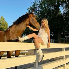 a woman sitting on top of a white fence next to a brown horse