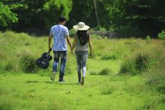 two people walking in the grass holding hands and carrying guitar cases, with trees in the background