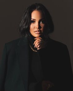 a woman wearing a black suit and gold necklace with her hand on her chin looking at the camera