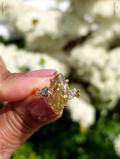 a hand holding a yellow diamond in front of some white flowered trees and bushes