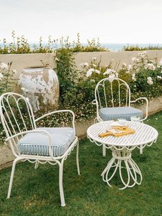 two white chairs and a table with food on it sitting in the grass near some flowers