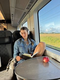 a woman sitting on a train reading a book and listening to headphones while looking out the window