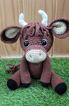 a crocheted cow sitting on top of green grass next to a wooden wall