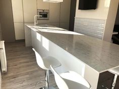 an empty kitchen with white stools and marble counter tops in front of a flat screen tv mounted on the wall