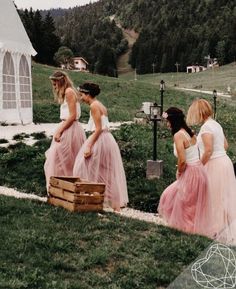 four women in long dresses are standing on the grass and looking at something behind them