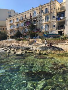 people are standing on the rocks by the water in front of an apartment building with several balconies