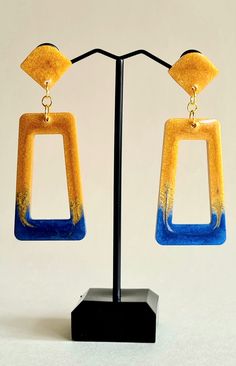 a pair of yellow and blue earrings on a black stand with a white wall in the background