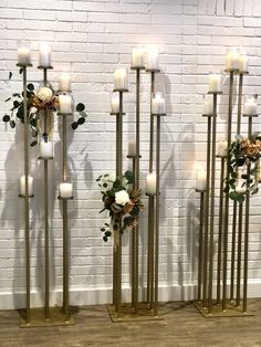 four tall metal candles with flowers and greenery on them in front of a white brick wall