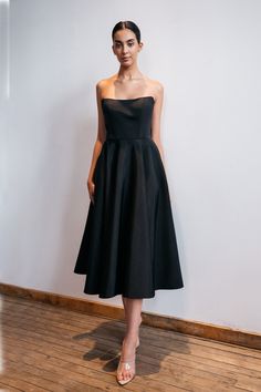 a woman standing in front of a white wall wearing a black strapless dress and heels