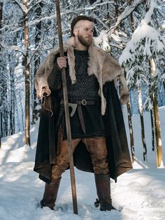 a man dressed in medieval clothing standing in the snow holding a stick and wearing a fur coat