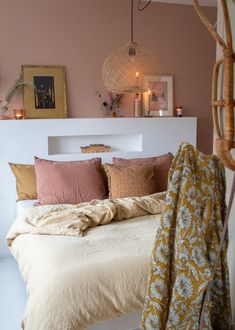 a bed with pillows and blankets on it in a room that has pink walls, wicker lights and pictures above the headboard