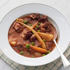 a white bowl filled with beef and potatoes on top of a table next to a napkin