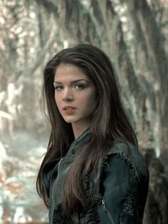 a woman with long hair standing in front of a tree filled forest and looking at the camera