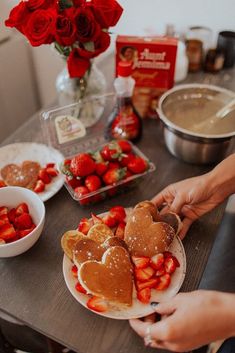 two people holding plates with heart shaped pancakes and strawberries on them next to bowls of fruit