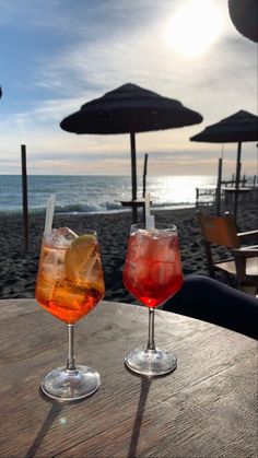 two drinks sitting on top of a wooden table next to the ocean with umbrellas in the background