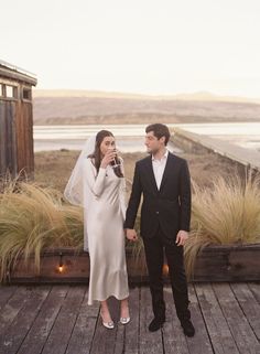 a bride and groom standing on a wooden deck holding wine glasses in front of them