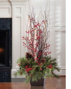 a vase filled with red berries and pine cones on top of a wooden floor next to a fireplace