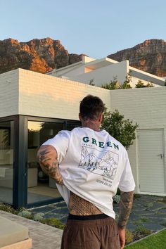 a man with tattoos standing in front of a house and looking at the mountains behind him