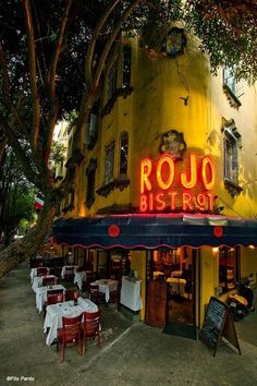 an outdoor restaurant with tables and chairs on the sidewalk next to it's neon sign