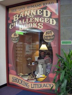 a window display with an advertisement for banned and challenged books in the front, behind which is a potted plant