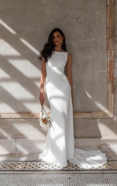 a woman standing in front of a wall wearing a white dress and holding a bouquet