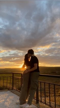 a man and woman standing on top of a balcony next to each other with the sun setting behind them