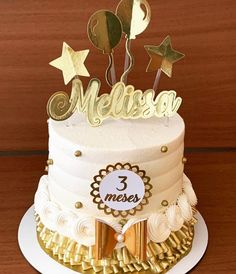 a three tiered cake with gold decorations and name written on the top, sitting on a wooden table