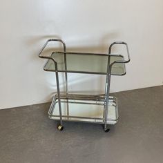 a metal and glass serving cart with wheels on the floor next to a white wall