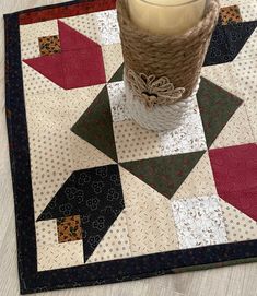 a candle is sitting on top of a quilted table runner with a vase in the middle