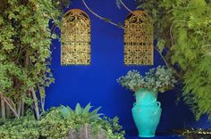 a blue wall with two gold bells hanging from it's sides and plants in the foreground