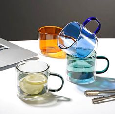 three mugs filled with different colored liquid next to a laptop on a white table