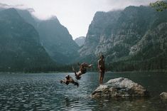 two people are jumping off rocks into the water in front of some mountains and trees